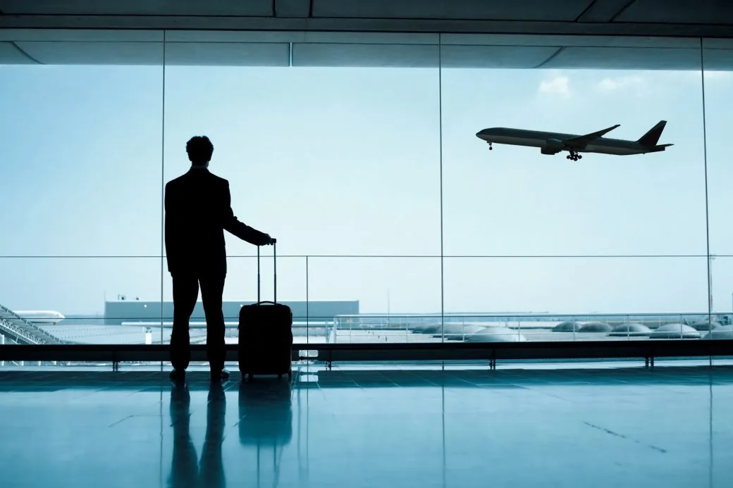 A traveller with a case at a departure lounge window, watching an aircraft take off.