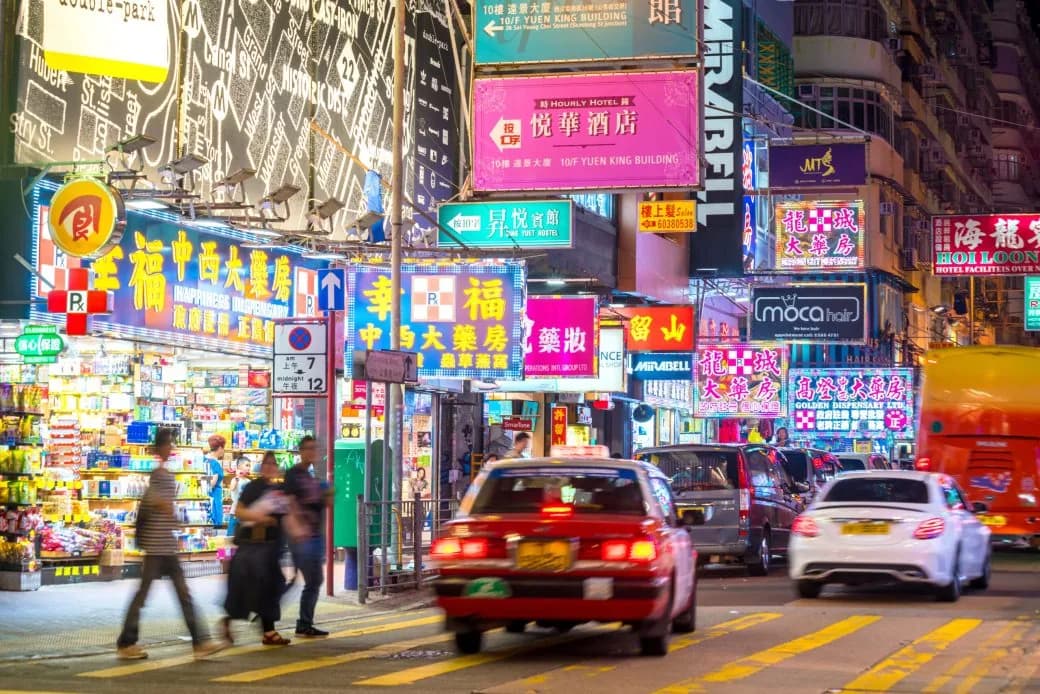 A neon-lit city street at night with shopfront signs, a red taxi and pedestrians.