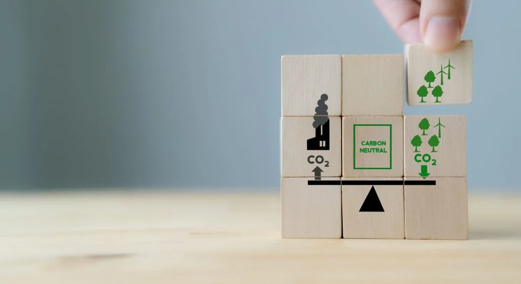 Wooden blocks balanced in a see-saw showing CO2 and carbon neutral symbols, with a hand adding trees and turbines.