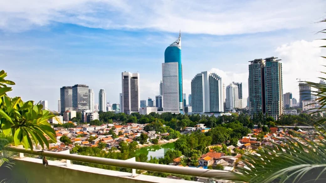 A city skyline with a tall glass tower rising above dense low-rise rooftops and palm trees in the foreground.