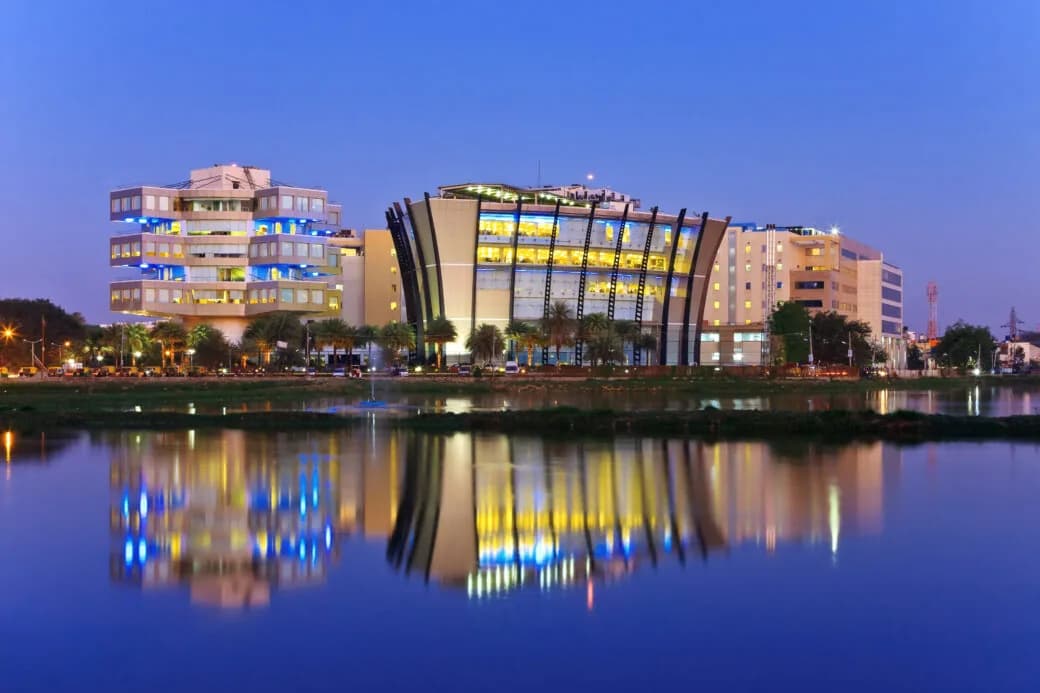 A modern glass office complex lit up at dusk, reflected in the water in front of it.