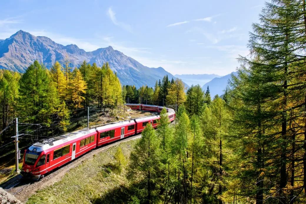 A red mountain train winding through pine forest with peaks rising behind it.