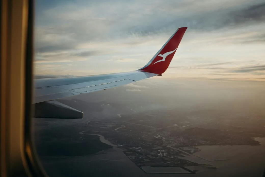 A Qantas aircraft wingtip seen from a window above a coastline at dusk.