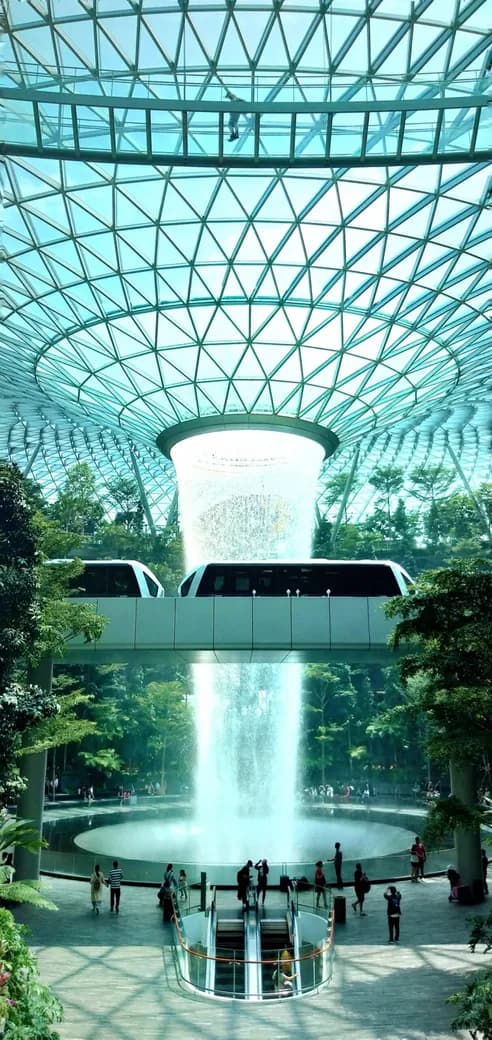 A monorail passes beneath a huge indoor waterfall inside a glass-domed airport atrium filled with trees.