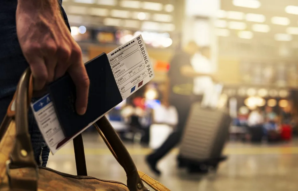 A traveller holds a passport and boarding pass while gripping a bag in an airport terminal.