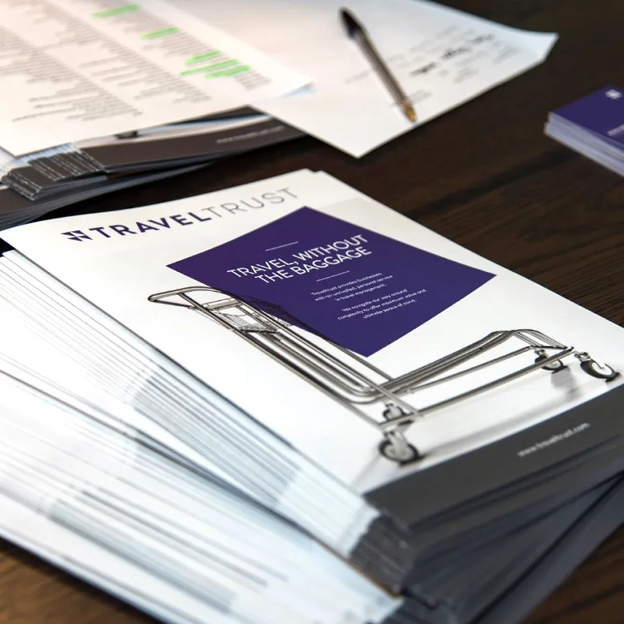 A stack of Traveltrust brochures reading Travel Without the Baggage on a table with papers and business cards.