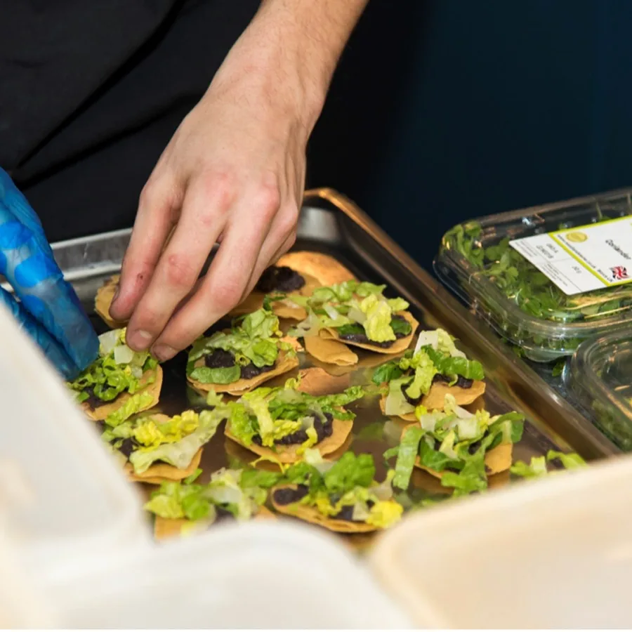 A gloved hand arranges garnished canapés on a tray beside a container of salad leaves.