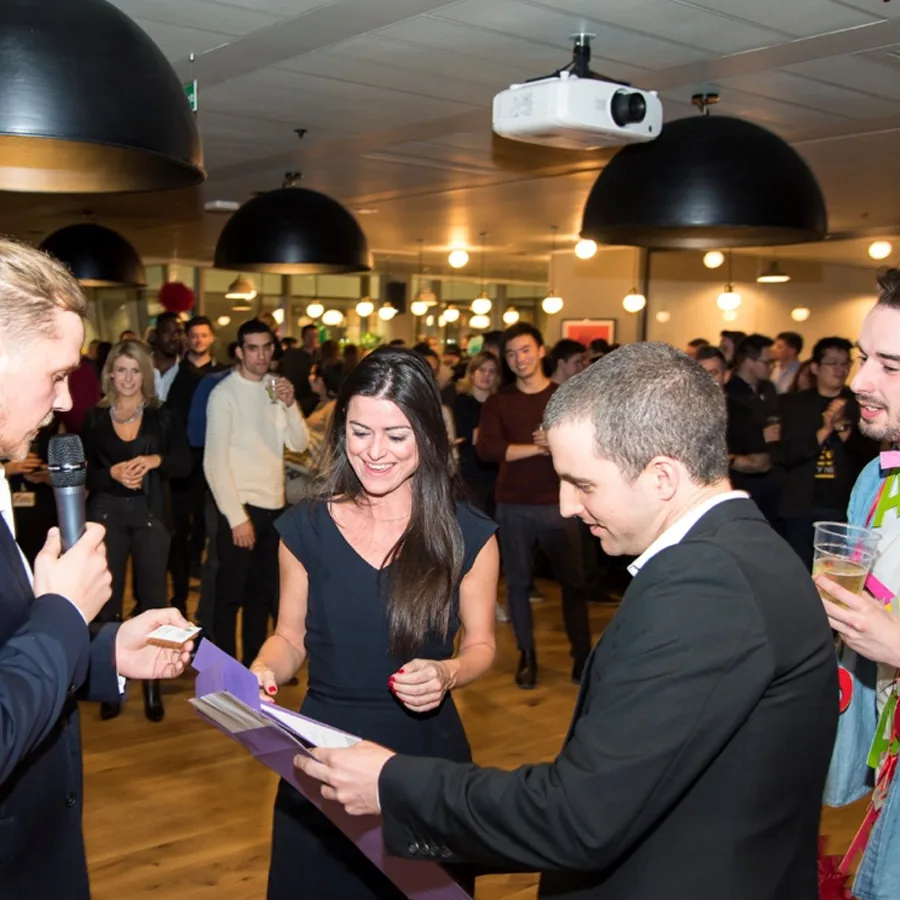 A crowd mingles at an evening reception as a man hands a card to a woman holding a folder.