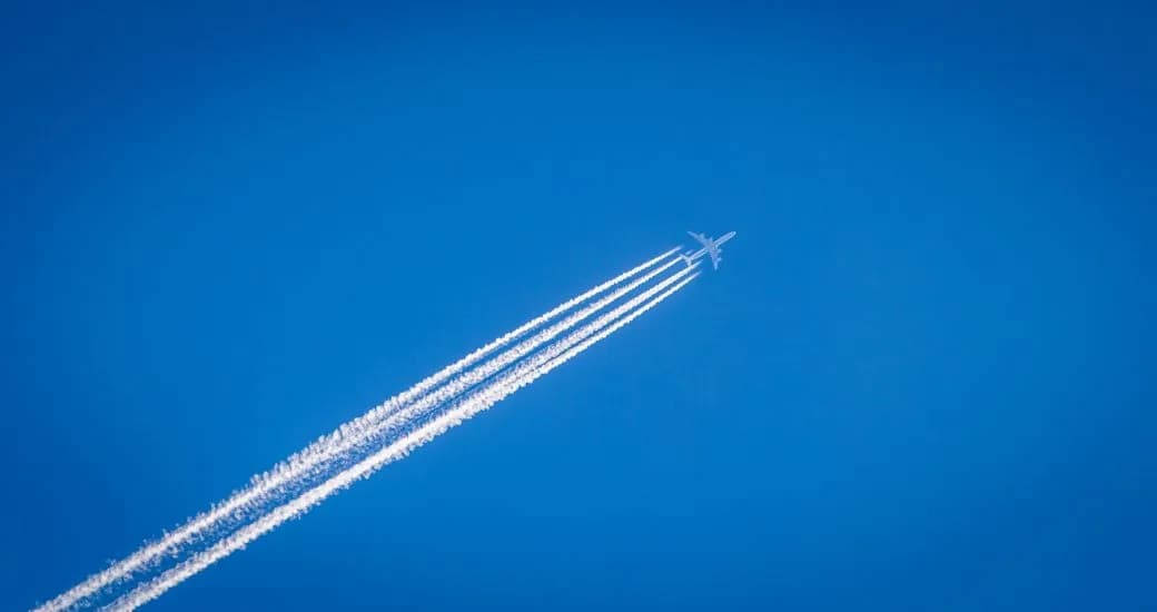 A distant aircraft leaves four contrails across a clear blue sky.