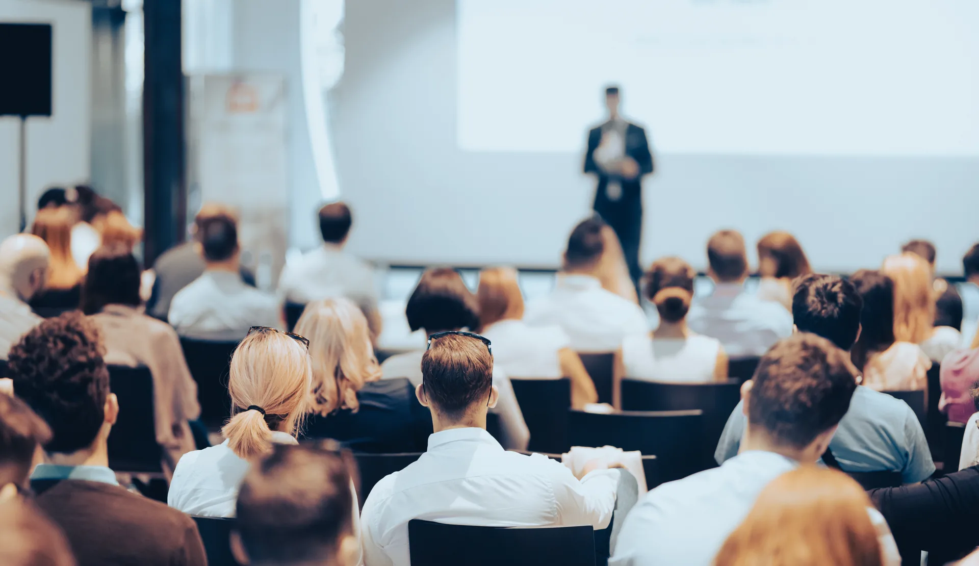 A conference audience seated in rows, listening to a speaker at the front of a bright hall.
