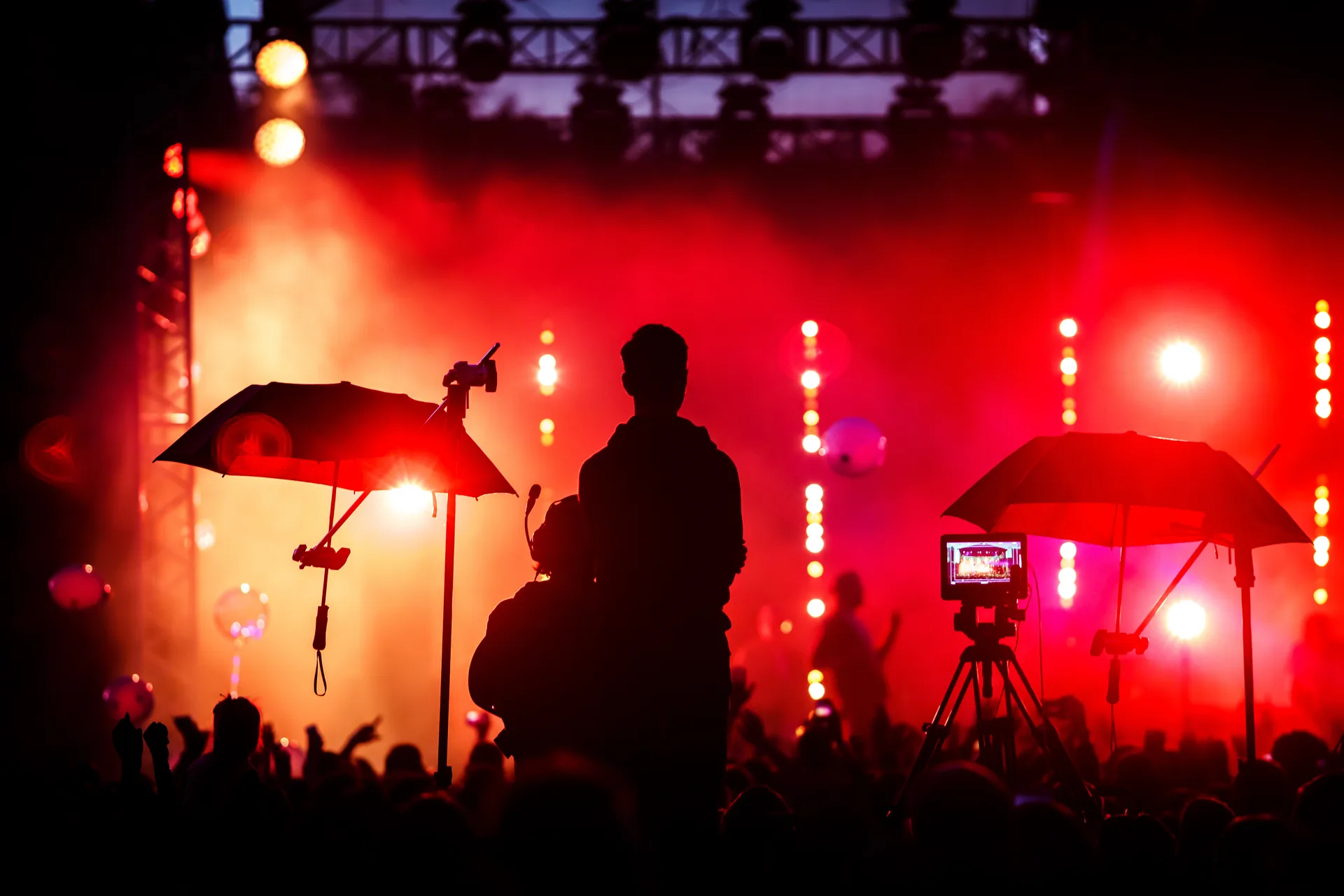 A crowd in front of a concert stage lit red, with a camera operator filming from the floor.