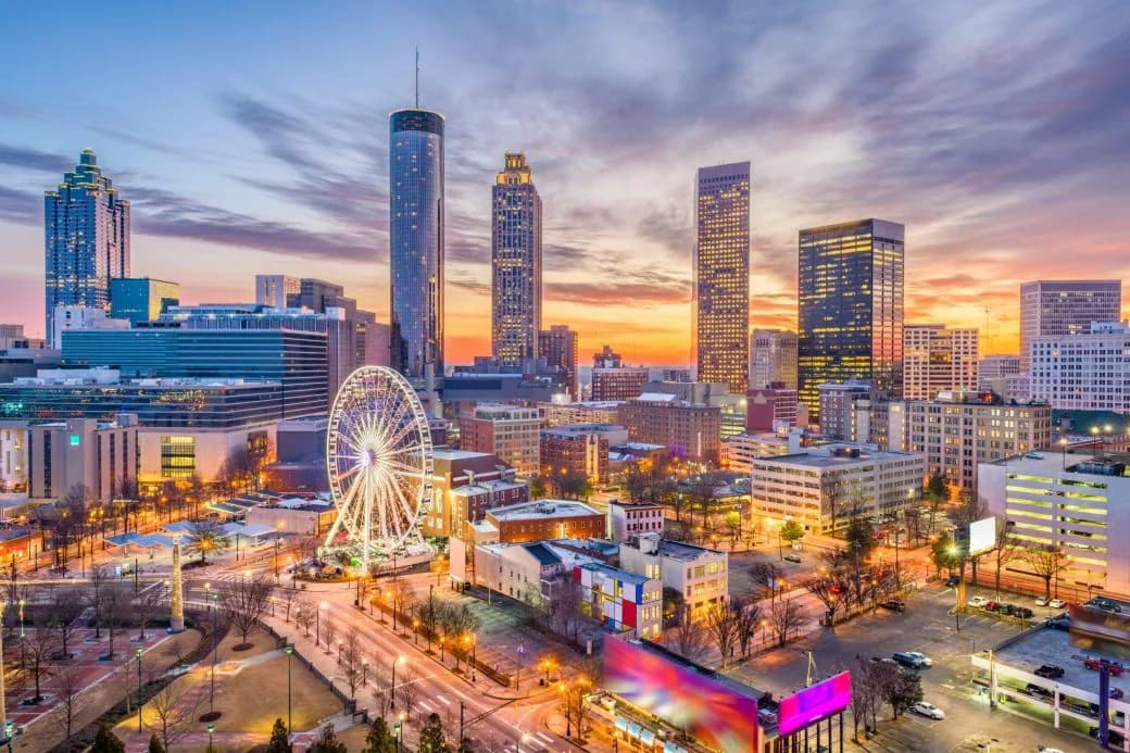 A city skyline at dusk with an illuminated ferris wheel and downtown towers.