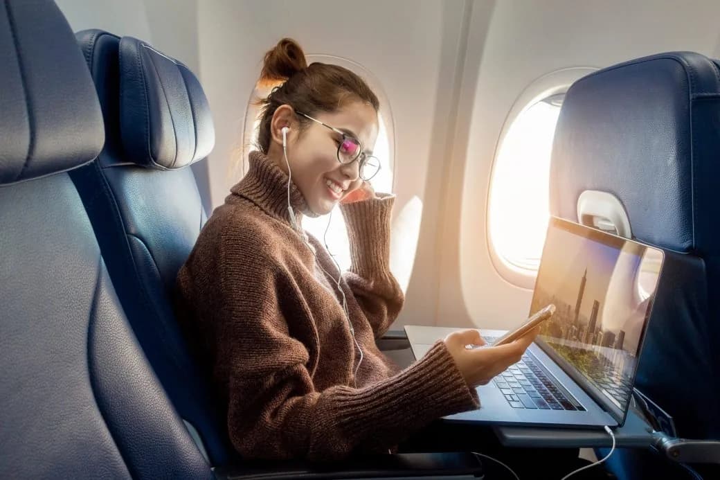 A smiling passenger using a laptop and phone in an aircraft window seat.
