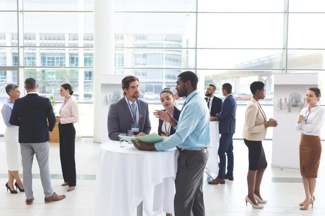Business people wearing name badges talking in a glass-walled conference lobby.