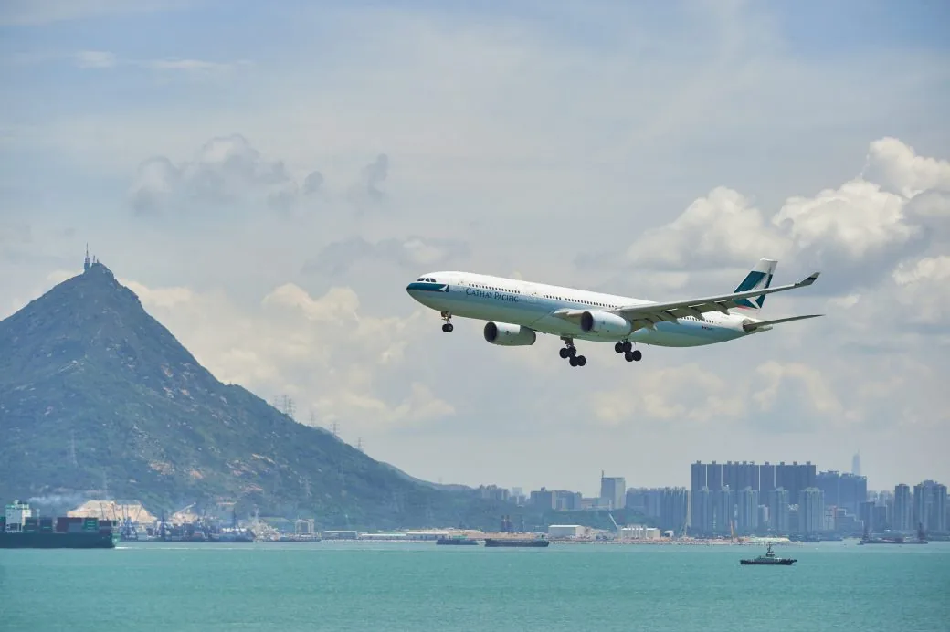 A Cathay Pacific aircraft descending over a harbour with a hill and container ships below.