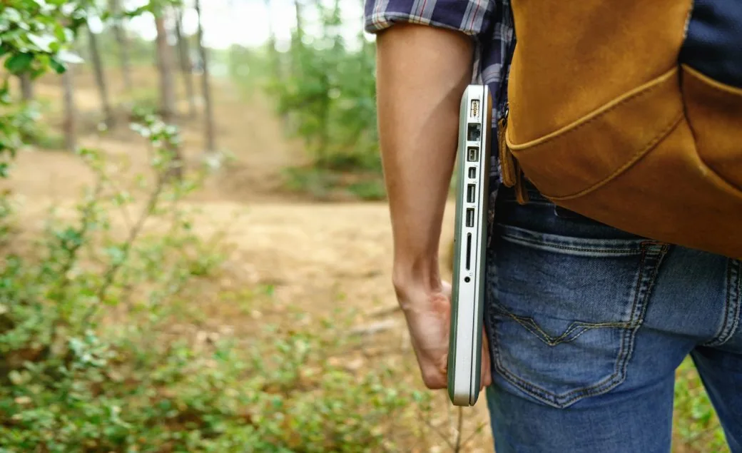 A person carrying a laptop under their arm while walking a wooded trail with a backpack.