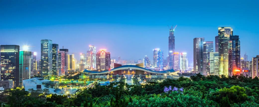 A city skyline lit up at dusk with colourful high-rise towers and greenery in the foreground.
