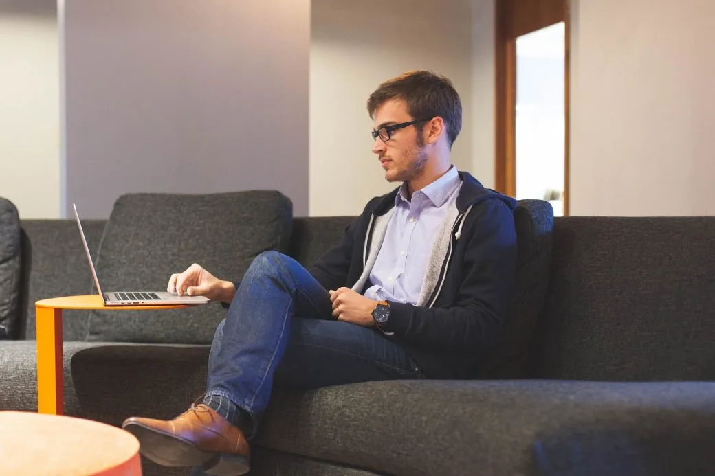 A man in glasses works on a laptop set on a low table while seated on a grey sofa.
