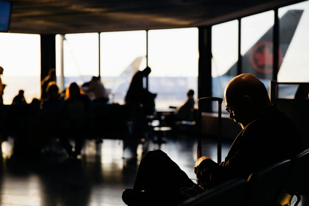 A traveller sits with a case in a dimly lit airport lounge as an Air Canada aircraft tail is visible through the window.