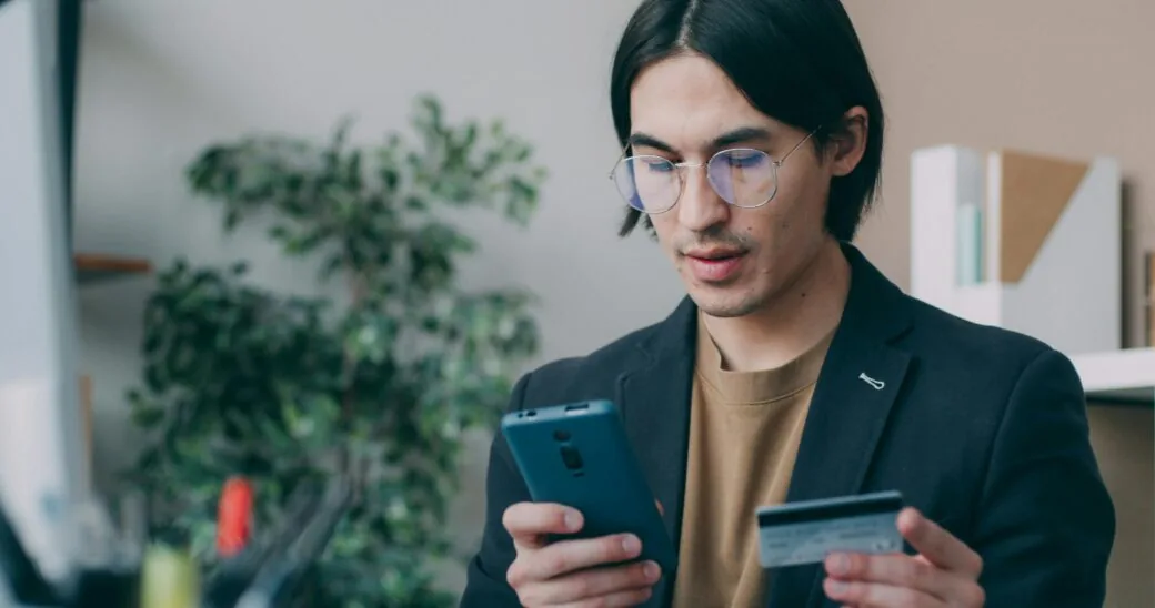 A man in glasses holds a phone and a card while standing indoors near a potted plant.