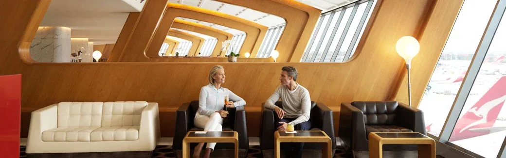 Two people chat over drinks in a wood-panelled airport lounge with a Qantas aircraft tail visible through the window.